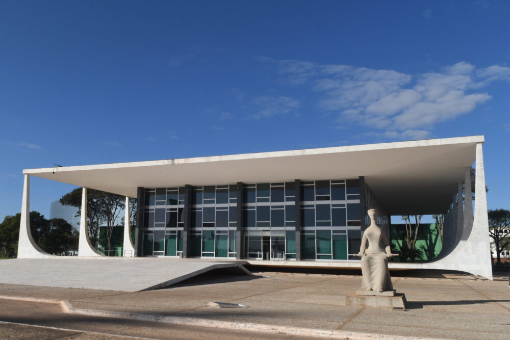Edifício-sede do STF, visto a partir da Praça dos Três Poderes, com a estátua da Justiça do lado direito da foto e a rampa que leva ao prédio do lado esquerdo. Luz do dia, céu azul.