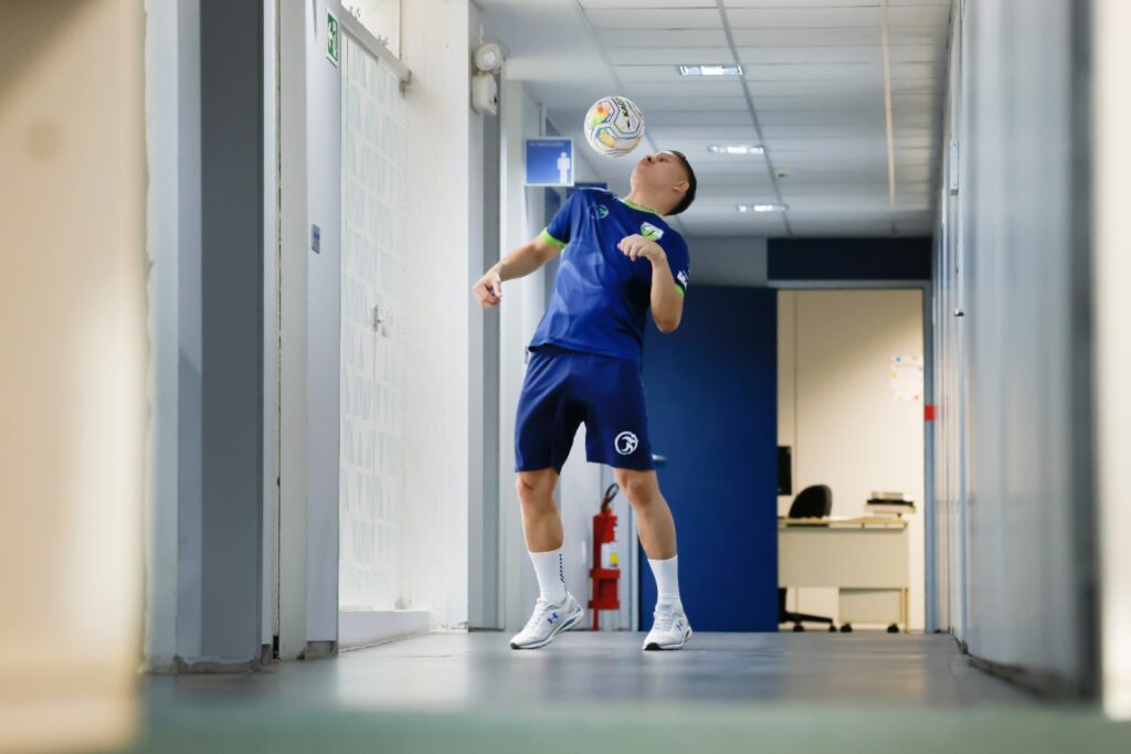 Foto colorida na horizontal mostra Ederson Rocha no corredor do local onde trabalha, no STF. Ele usa o ombro para fazer um movimento com a bola de futsal. Veste uniforme azul e meias brancas.