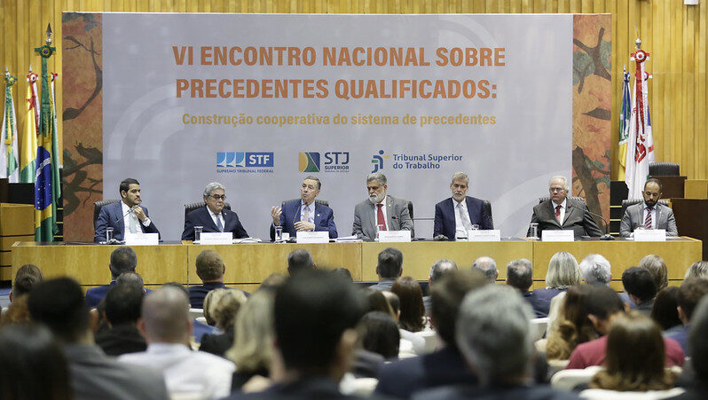 Foto colorida na posição retrato mostra mesa de palestrantes do VI Encontro Nacional sobre Precedentes Qualificados. Ao fundo, painel com o nome do evento. Logo à frente do painel estão sete palestrantes sentados. Entre eles, no centro, está o presidente do STF, que faz um gesto com as mãos enquanto fala. No primeiro plano da foto, desfocado, vemos o público sentado no auditório.