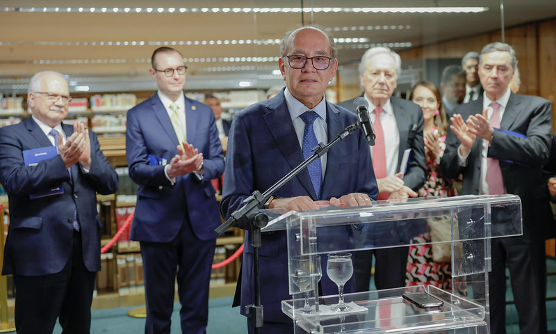 Foto colorida no formato paisagem do ministro Gilmar Mendes no púlpito em frente à bibliotrca do STF. Atrás dele estão os ministros Barroso, Zanin e Fachin e o ministro aposentado Lewandoviski.