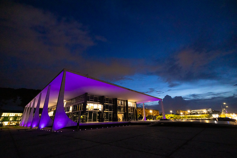 Foto colorida na horizontal do edifício-sede do STF iluminado na cor roxa. Foto noturna.