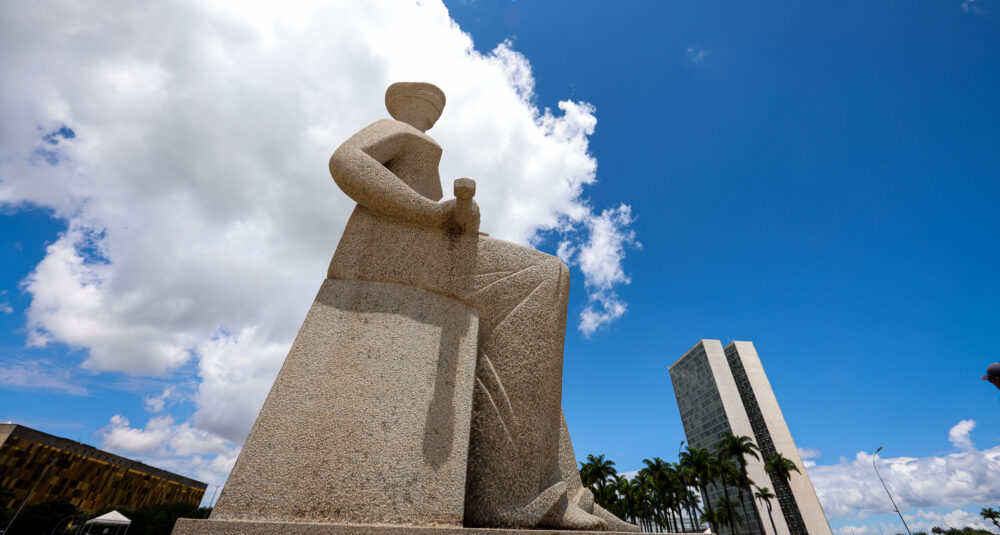 Foto em formato paisagem da estátua da Justiça de lado. A esquerda o prédio da Câmara dos Deputados e a direita o prédio do Congresso Nacional com palmeiras e o fundo céu azul