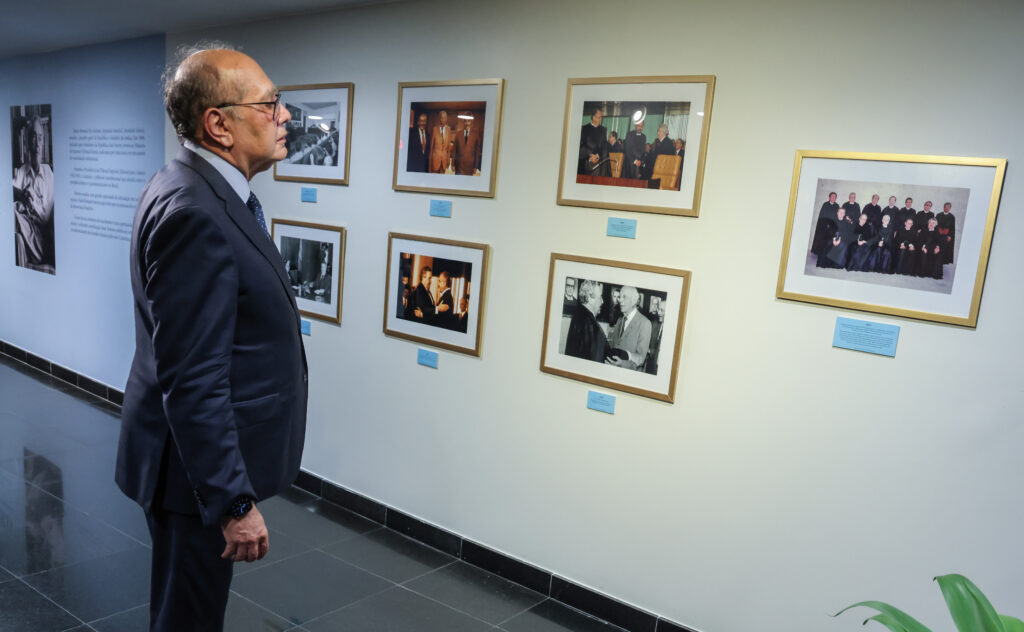 Foto colorida na horizontal do ministro Gilmar Mendes diante de quadros com fotografias na parede.