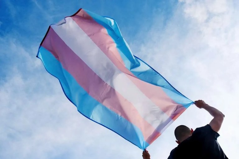 Detalhe de pessoa segurando a bandeira da visibilidade trans, com faixas horizontais azuis, rosa e brancas