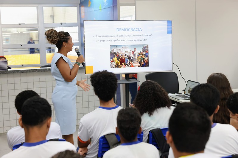 Foto colorida na horizontal com a juíza Flávia do lado esquerdo. Ela é uma mulher negra. Na sua frente um telão onde está escrito democracia. Abaixo tem um texto e uma foto. Diante do telão aparecem jovens sentados em carteiras olhando pra a tela.
