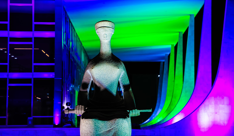 Foto colorida da fachada do STF iluminada com as cores da campanha do Dia Internacional da Mulher (roxo, verde e branco). Em primeiro plano, na foto, aparece a estátua da justiça feita de material sólido, com vendas nos olhos e espada deitada sobre as pernas, A estátua está em posição sentada.