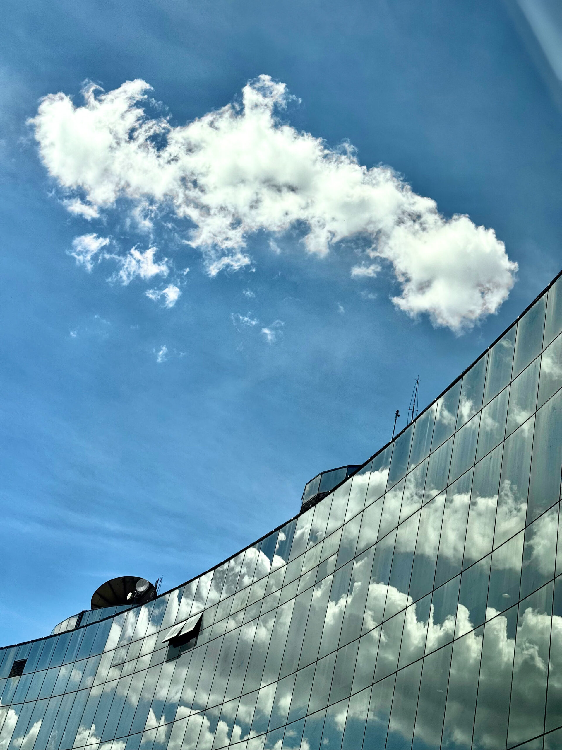 A imagem mostra a parte superior da fachada do STF em formato curva e espelhada de vidro, refletindo tons suaves do céu. O fundo exibe um céu azul claro com nuvens esbranquiçadas.