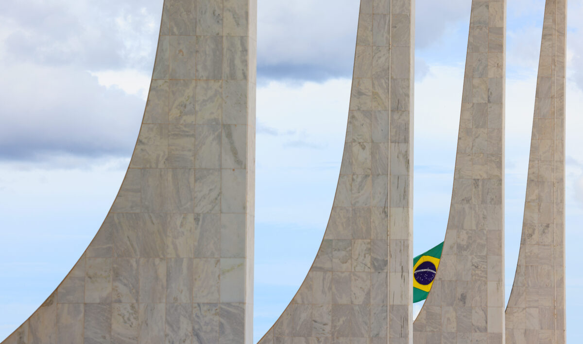 A imagem mostra colunas de mármore curvadas, do prédio do STF formando parte de uma estrutura arquitetônica moderna. Entre os espaços vazios das colunas, é possível ver ao fundo uma bandeira do Brasil tremulando, sob um céu parcialmente nublado.
