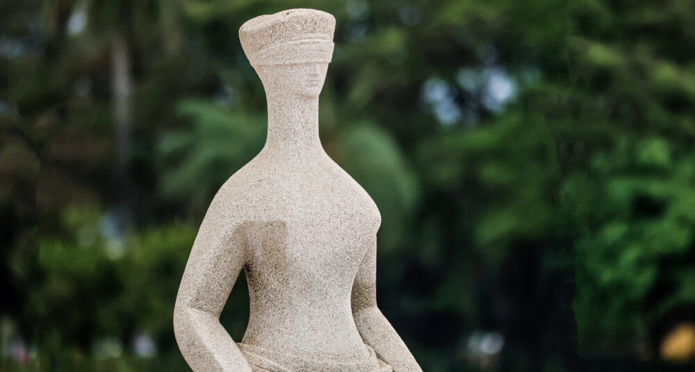 Foto colorida na horizontal da estátua da justiça que tem vendas nos olhos e uma espada deitada no colo. Ao fundo, desfocado, aparece o verde de árvores