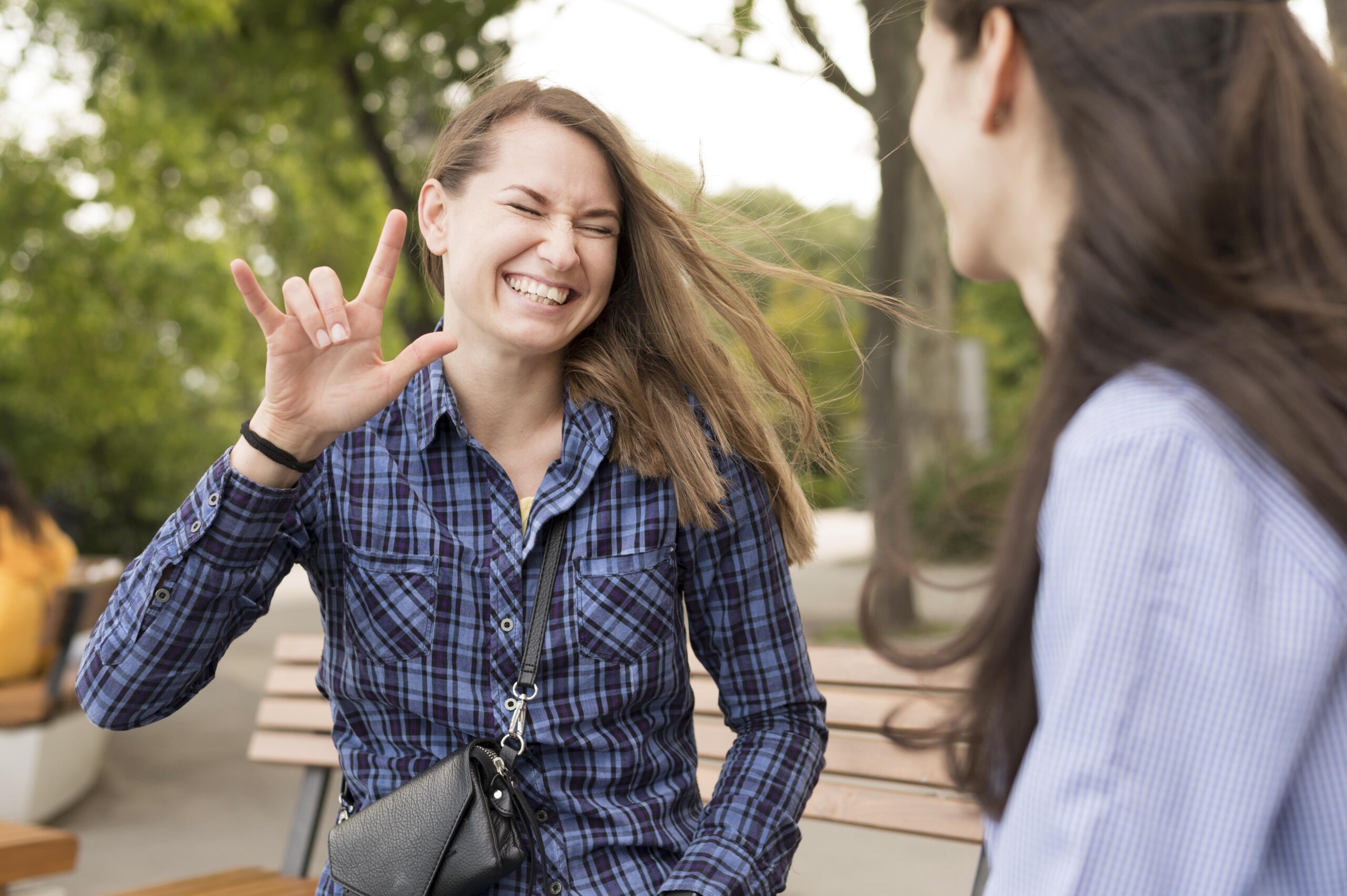 Duas mulheres estão sentadas em um banco ao ar livre. Uma delas sorri e faz o sinal de 