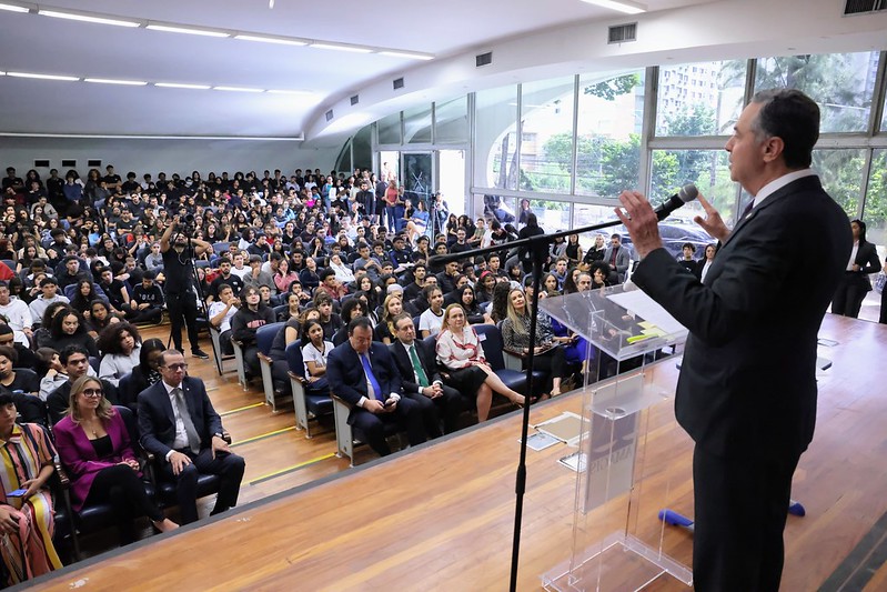 Fotografia colorida, na posição horizontal, de uma auditório lotado com o ministro Luís Roberto Barroso posicionado à direita, em um púlpito, proferindo a palestra