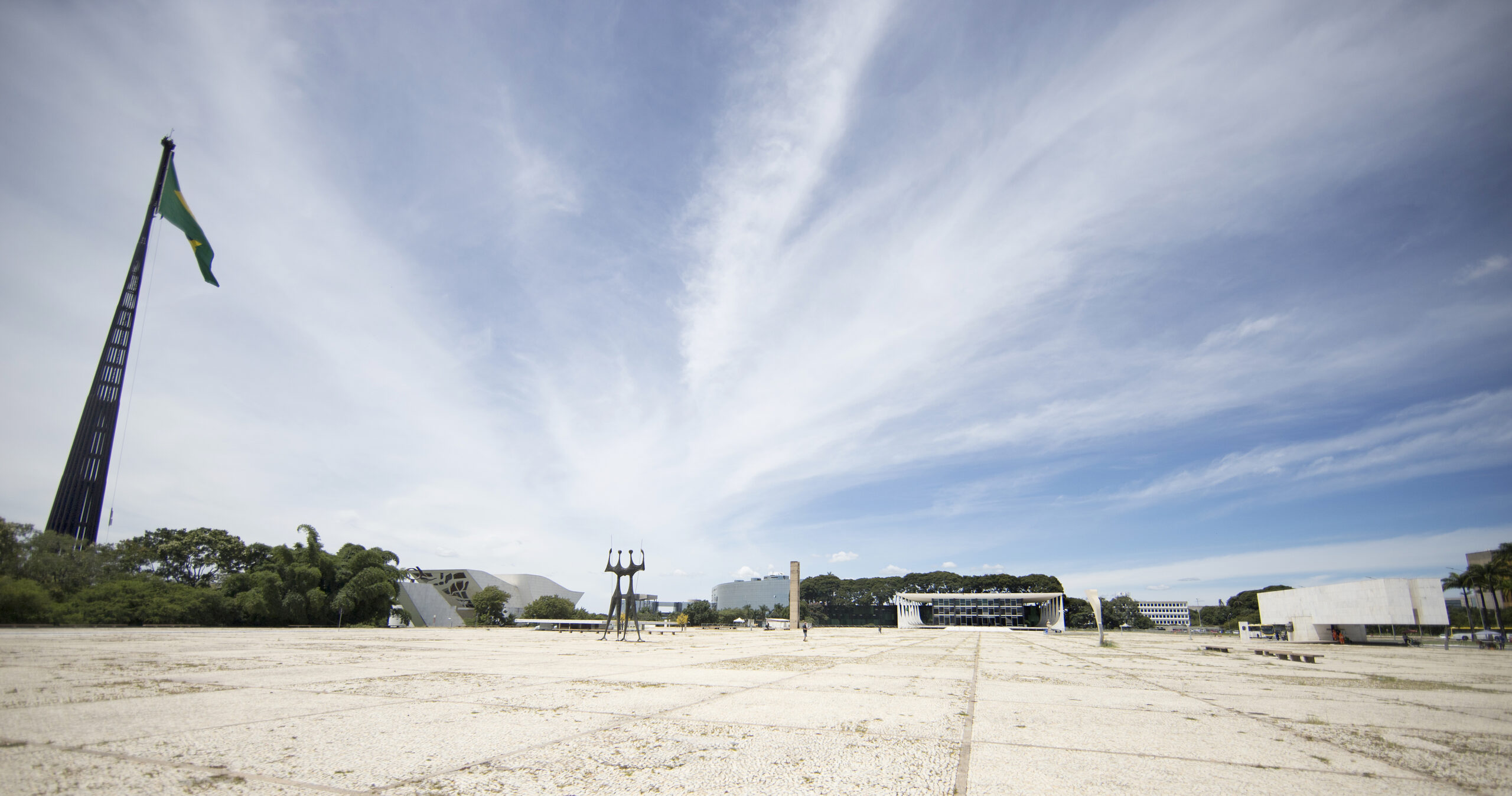Foto colorida em panorâmica da Praça dos 3 poderes, do lado esquerdo a bandeira do brasil, em seguida o monumento aos candangos, depois o prédio do STF. O céu é azul claro e com nuvens que formam uma névoa fina.