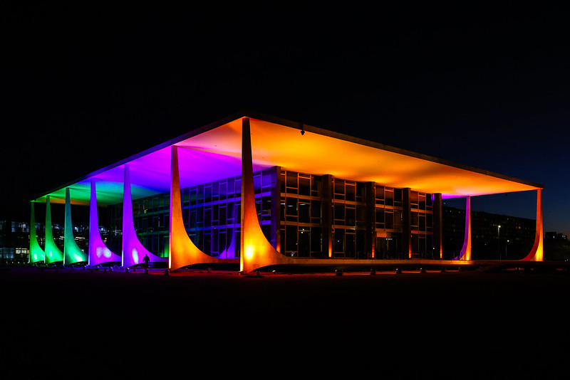 Foto noturna da Fachada do STF iluminada nas cores laranja, roxo e verde.