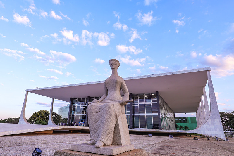 Foto colorida da estátua da Justiça com o edifício-sede do STF ao fundo. O ângulo é de baixo para cima, o céu aparece em tom azul claro e com algumas nuvens espalhadas