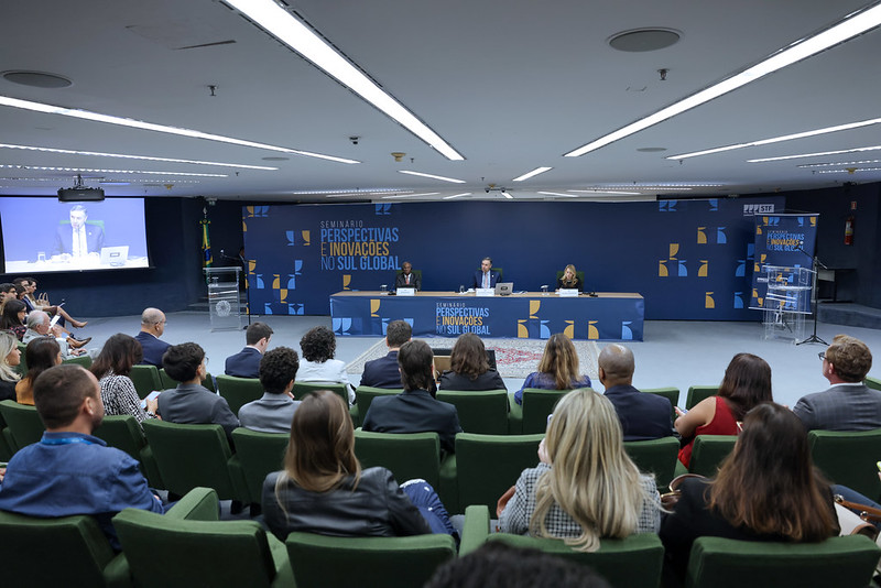 Fotografia do evento. Ministro Barroso na mesa ladeado por dois convidados e de frente ao auditório lotado.