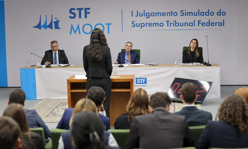Aluna de costas fazendo sustentação oral no púlpito perante a mesa onde estão o ministro Flávio Dino, o ministro Barroso ao centro e assessora do Plenário.