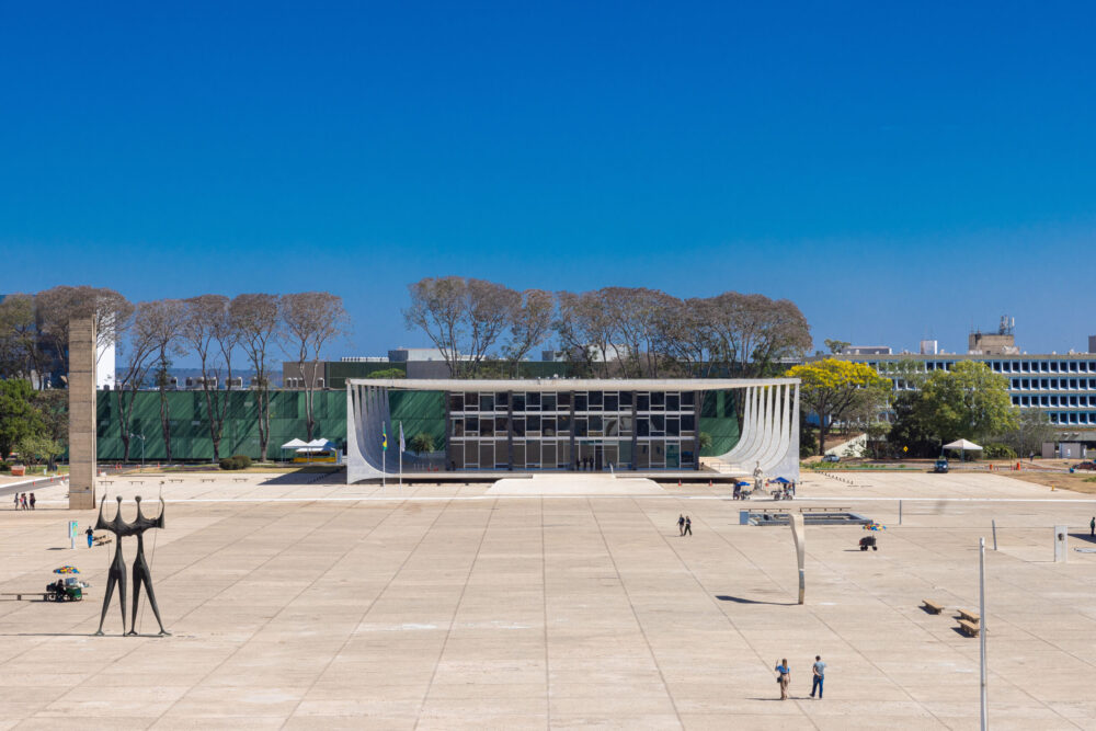 A imagem mostra a fachada principal do Supremo Tribunal Federal, em Brasília, sob um céu azul e sem nuvens. O prédio, de arquitetura moderna de Oscar Niemeyer, tem grandes colunas curvas nas laterais e uma ampla fachada de vidro. Em frente, estende-se uma esplanada clara e ampla, com poucas pessoas caminhando. À esquerda, destaca-se o monumento “Os Candangos”, escultura de bronze de Bruno Giorgi, com duas figuras humanas altas e alongadas, de braços erguidos e unidos no alto, simbolizando força e união. Atrás do edifício, há árvores de galhos finos e construções modernistas, compondo o cenário característico da capital federal.