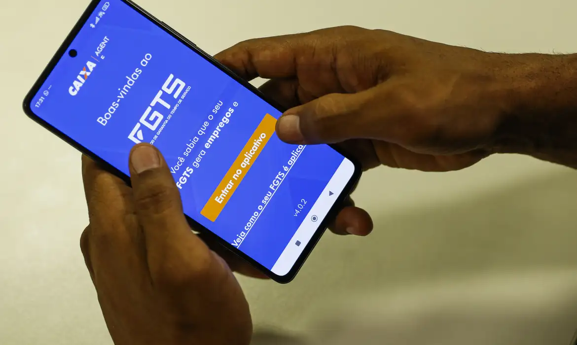 Fotografia colorida das mãos de uma pessoa negra segurando um celular, e na tela do aparelho aparece o aplicativo do FGTS.