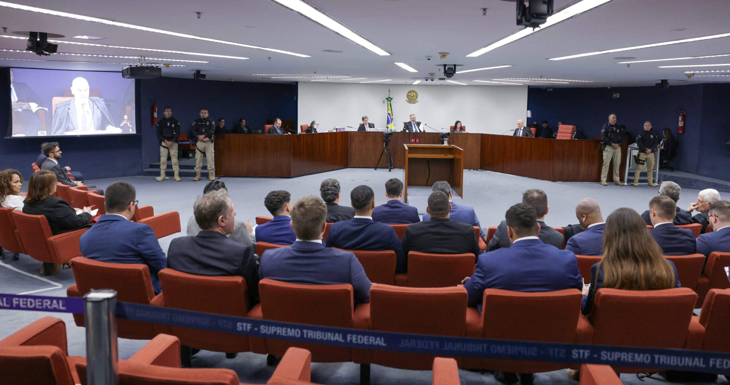 A imagem mostra a sala de sessões da Primeira Turma do Supremo Tribunal Federal durante o julgamento da Ação Penal 2696. No centro, há uma mesa em formato semicircular onde estão sentados os ministros e o procurador-geral da República com o brasão da República e a bandeira do Brasil ao fundo. À frente, ficam as cadeiras do público, ocupadas por homens e mulheres vestidos formalmente. Guardas de segurança, com uniformes escuros e calças bege, estão posicionados nas laterais. Um telão exibe a imagem ampliada do ministro Alexandre de Moraes falando. O ambiente é iluminado por luz branca, com paredes azuis e cadeiras de cor vermelha.