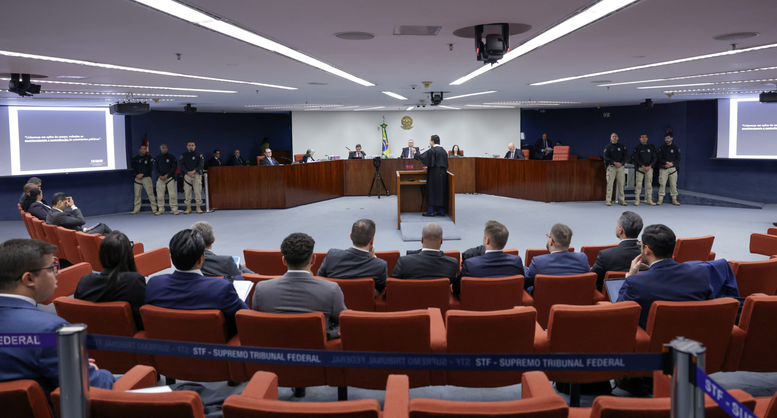 A imagem mostra a sala de sessões da Primeira Turma do Supremo Tribunal Federal durante o julgamento da Ação Penal 2696, do núcleo 3 da tentativa de golpe de estado. Ao centro, um advogado de beca preta fala em pé, de frente para os ministros e o representante da Procuradoria-Geral da República, que estão sentados atrás de uma grande bancada semicircular de madeira. Atrás deles, na parede branca, há o brasão da República e a bandeira do Brasil. Nas laterais, quatro agentes de segurança, com uniformes pretos e calças bege, permanecem de pé. À frente, o público assiste sentado em poltronas vermelhas, voltado para os ministros. Duas telas laterais exibem trechos de texto em fundo escuro. O ambiente é iluminado por luzes brancas embutidas no teto.