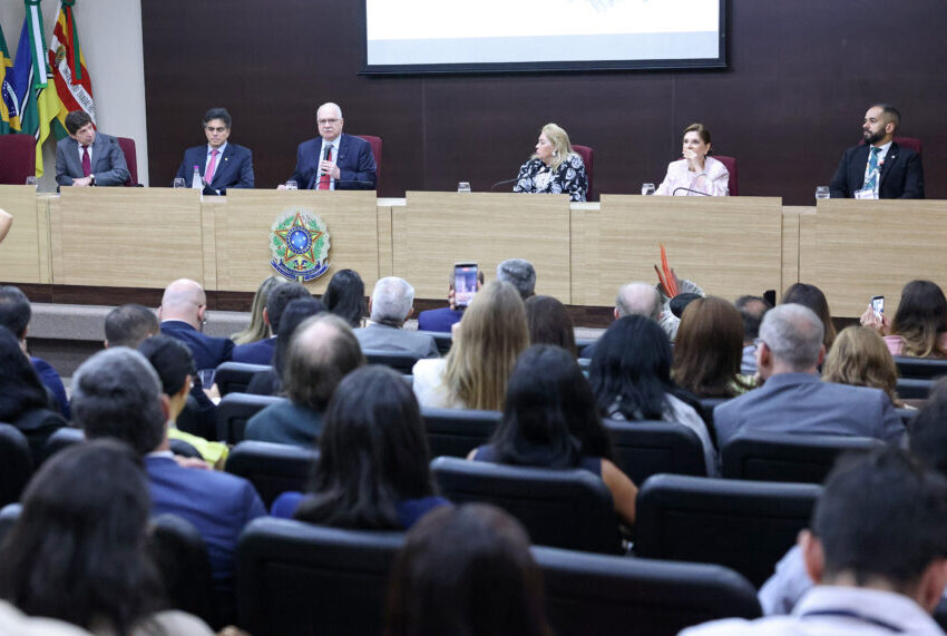 A imagem mostra o auditório do Tribunal Regional do Pará, na cidade de Belém, com dezenas de pessoas sentadas de frente para uma mesa elevada onde ocorre a abertura da 2ª Conferência Internacional de Sustentabilidade no Poder Judiciário, na COP 30. Na mesa, ao centro, o presidente do Supremo Tribunal Federal, ministro Edson Fachin, e mais seis participantes, três mulheres e três homens, sentados lado a lado, cada um com microfone e copo de água. Atrás deles, bandeiras do Brasil, de estados e de instituições estão posicionadas no canto esquerdo. Um telão ao fundo exibe parte da identidade visual do evento. No público, várias pessoas registram a cena com celulares. A sala tem cadeiras organizadas em fileiras e iluminação uniforme.