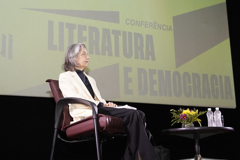 Fotografia da ministra Cármen Lúcia sentada e, atrás dela, um telão com o nome do evento 