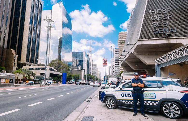Fotografia da avenida Paulista, na altura do prédio da Fiesp, com detalhe do lado direto para uma viatura da GCM e um agente da corporação.
