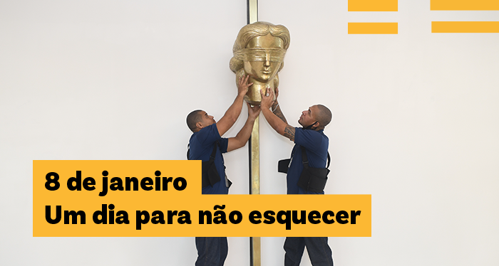 Fotografia de dois homens colocando na parede uma escultura que representa a cabeça da estátua da Justiça. No canto inferior esquerdo, está escrito o texto 