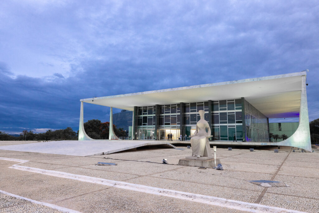 Foto em formato paisagem do prédio do STF ao entardecer. Estátua da Justiça em primeiro plano.