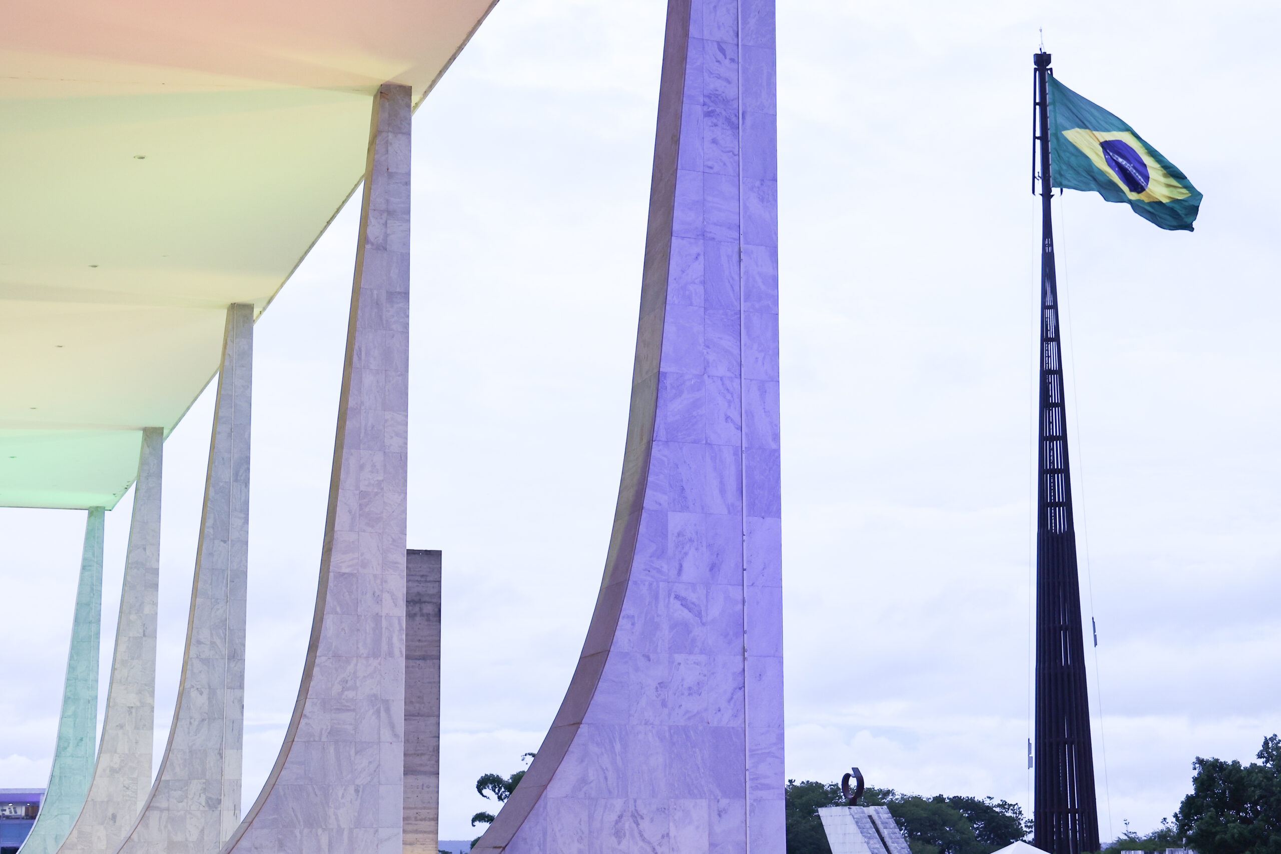 Foto dos arcos do prédio do STF, à esquerda. À direita, a Bandeira do Brasil