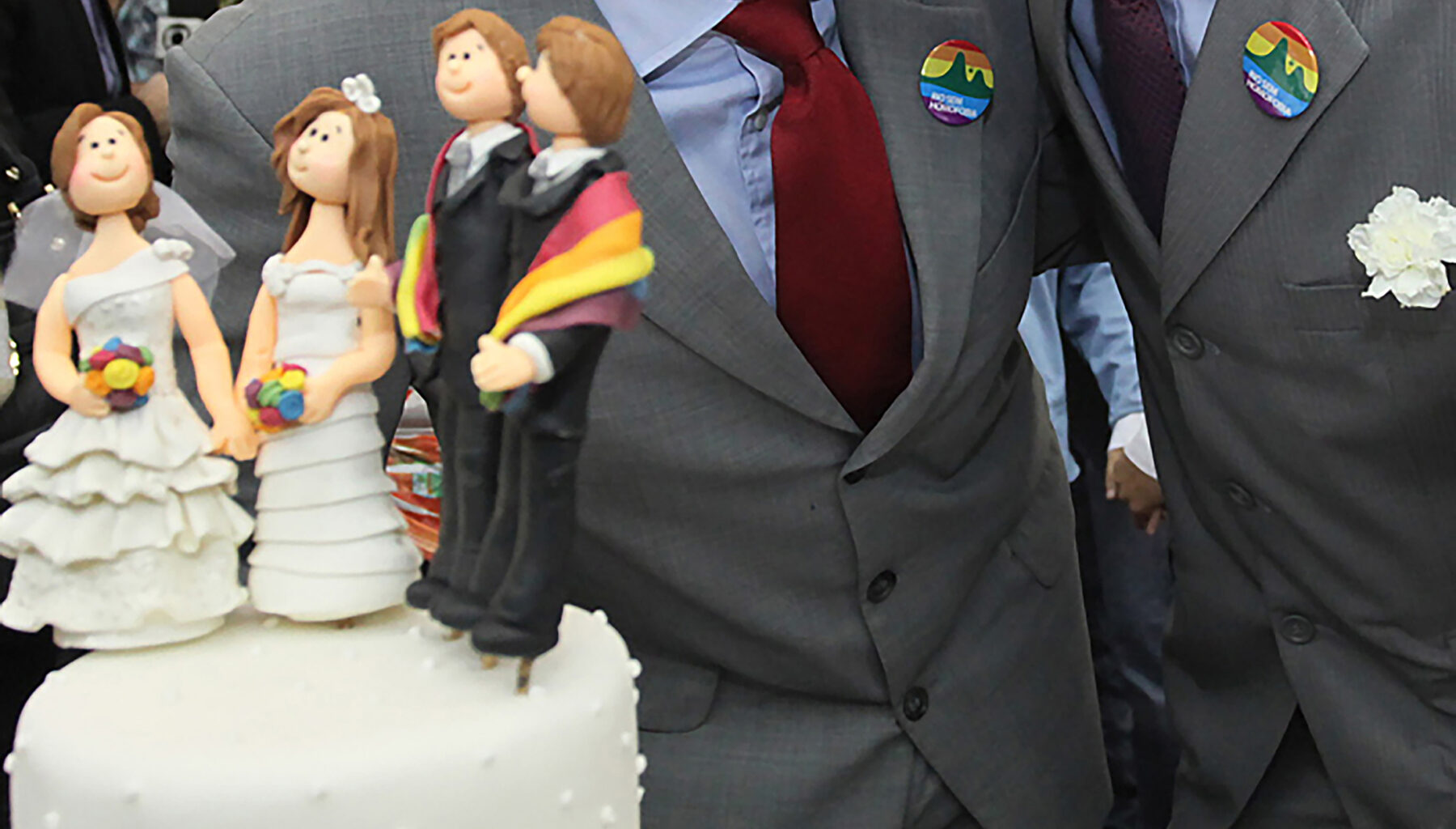 Foto de um bolo de casamento com quatro bonecos representando duas noivas e dois noivos, com duas pessoas atrás. Todos possuem adornos com os núcleos da bandeira do arco-íris, principal símbolo do movimento LGBTQIA+