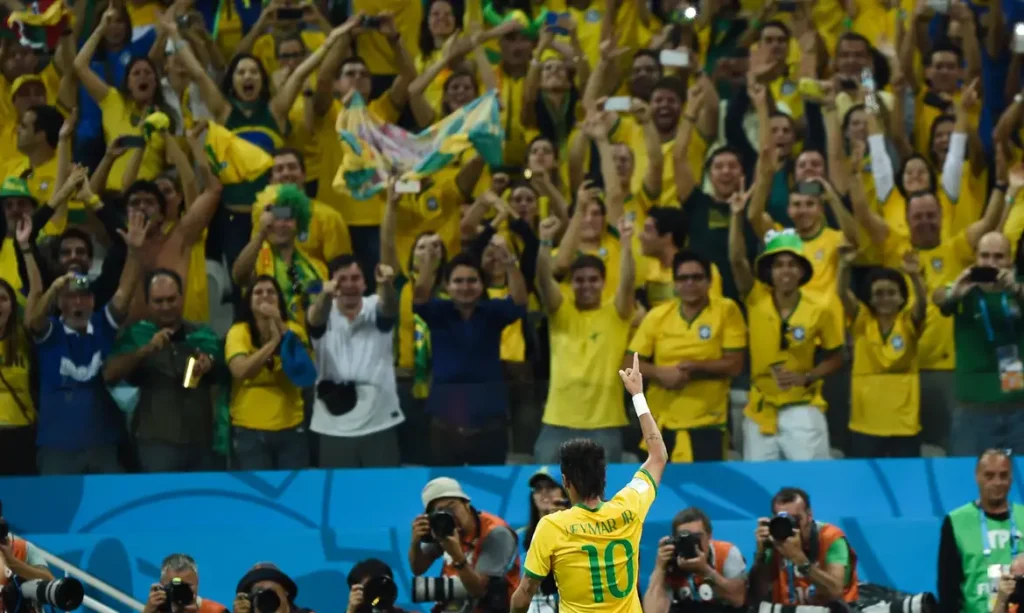 Fotografia com jogador Neymar Jr de frente para a torcida. Todos de mãos para cima comemorando.