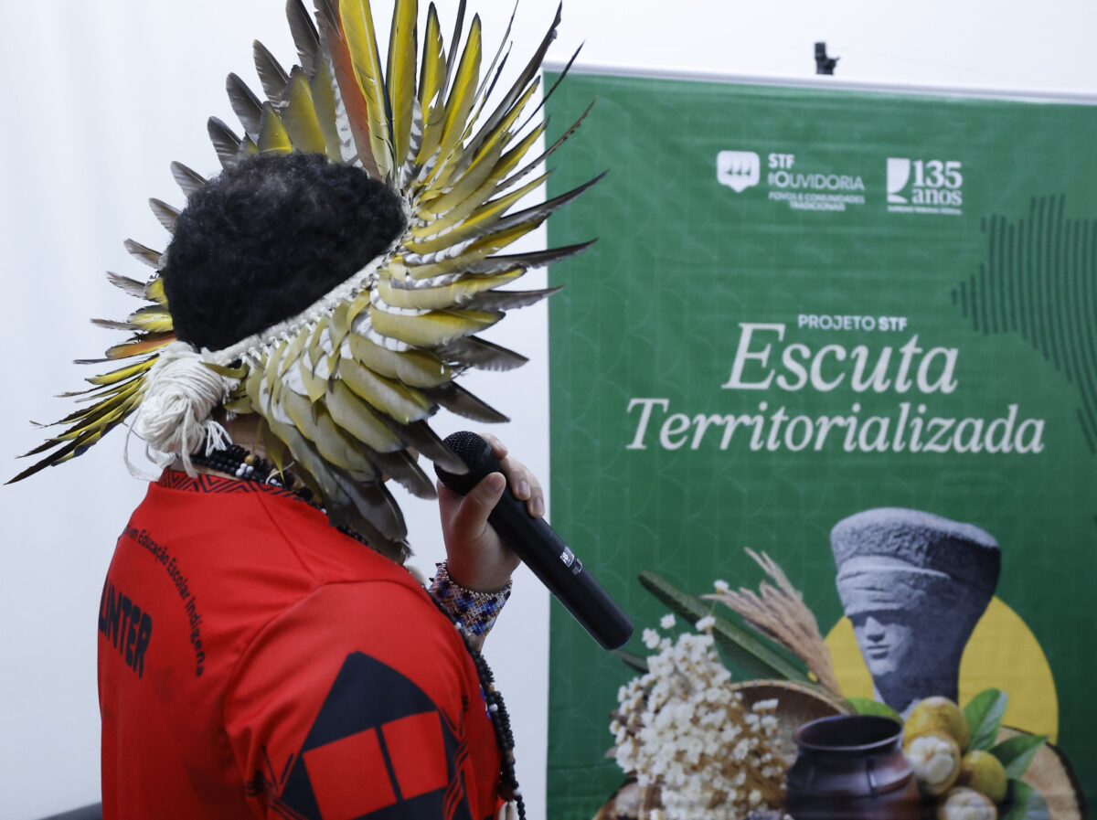 Fotografia de uma indígena de lado falando com um microfone na mao. Ao seu lado, banner do projeto Escuta Territorializada.