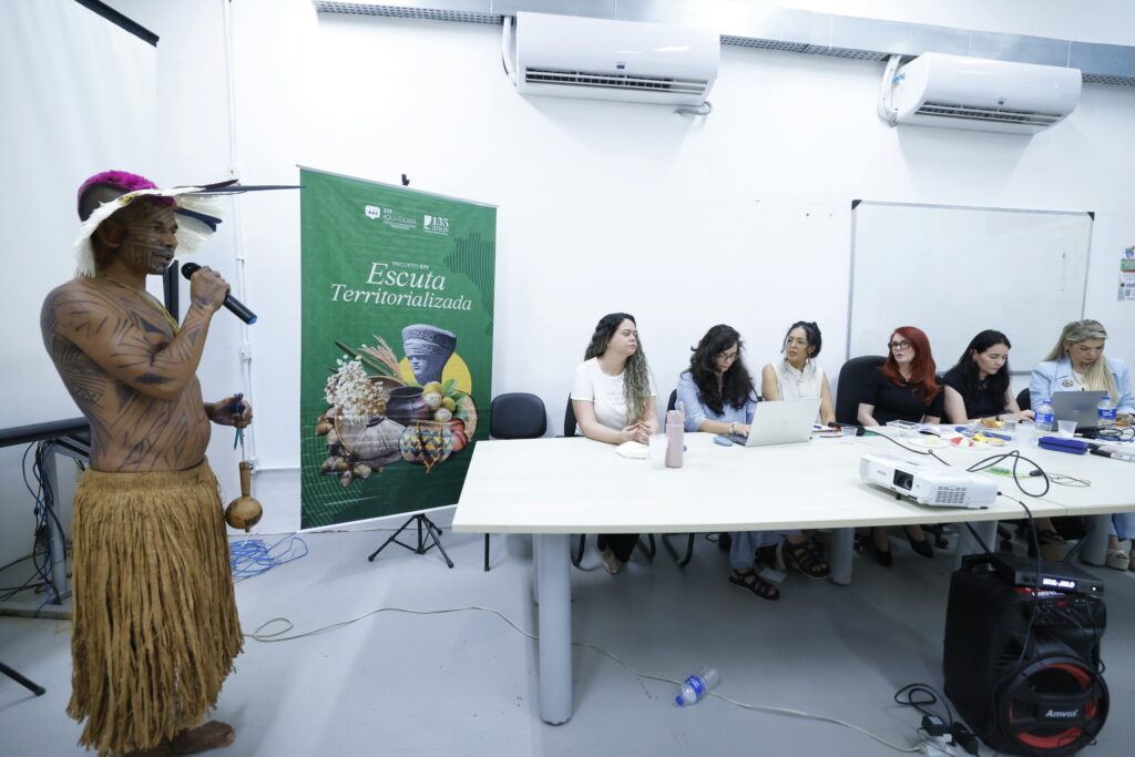 Fotografia de um indígena falando para as pessoas que compõem a mesa de escuta.
