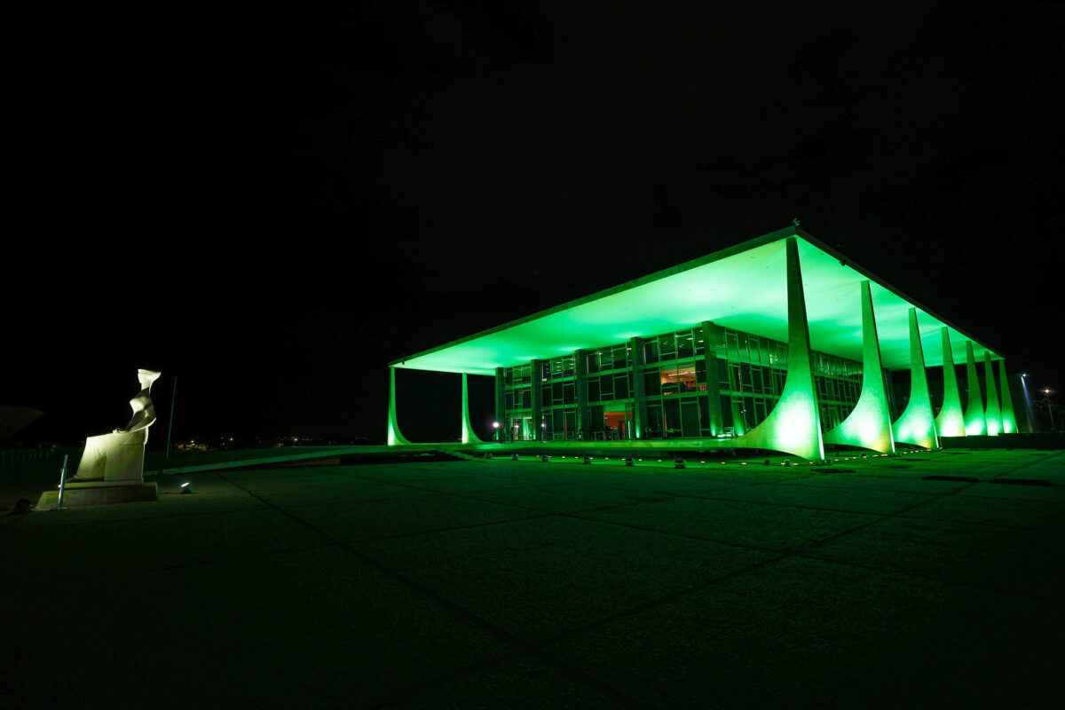 Fotografia do edifício-sede do STF iluminado de verde. Do lado esquerdo, a estátua da Justiça.