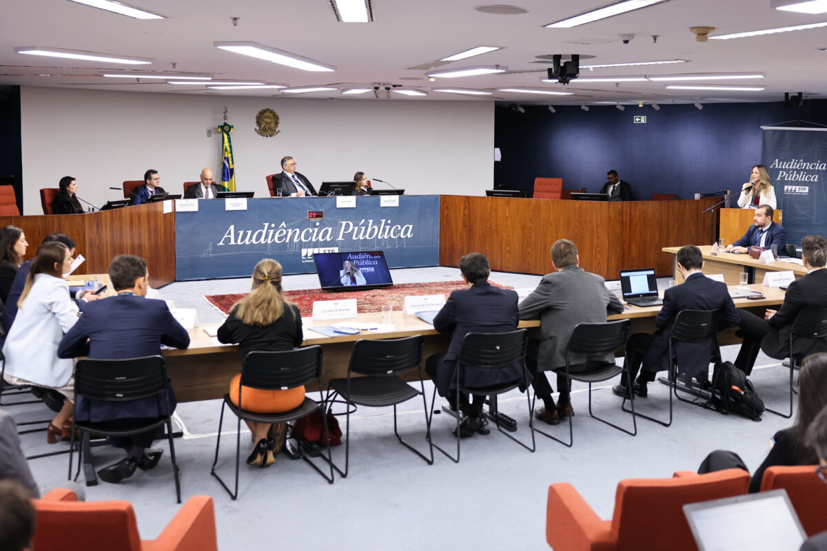 Fotografia da sala de sessões da Primeira Turma do STF em audiência pública. No centro da bancada, o ministro Flávio Dino. Do lado esquerdo, o ministro Alexandre de Moares. À direta, no púlpito, uma mulher loira faz sua exposição.