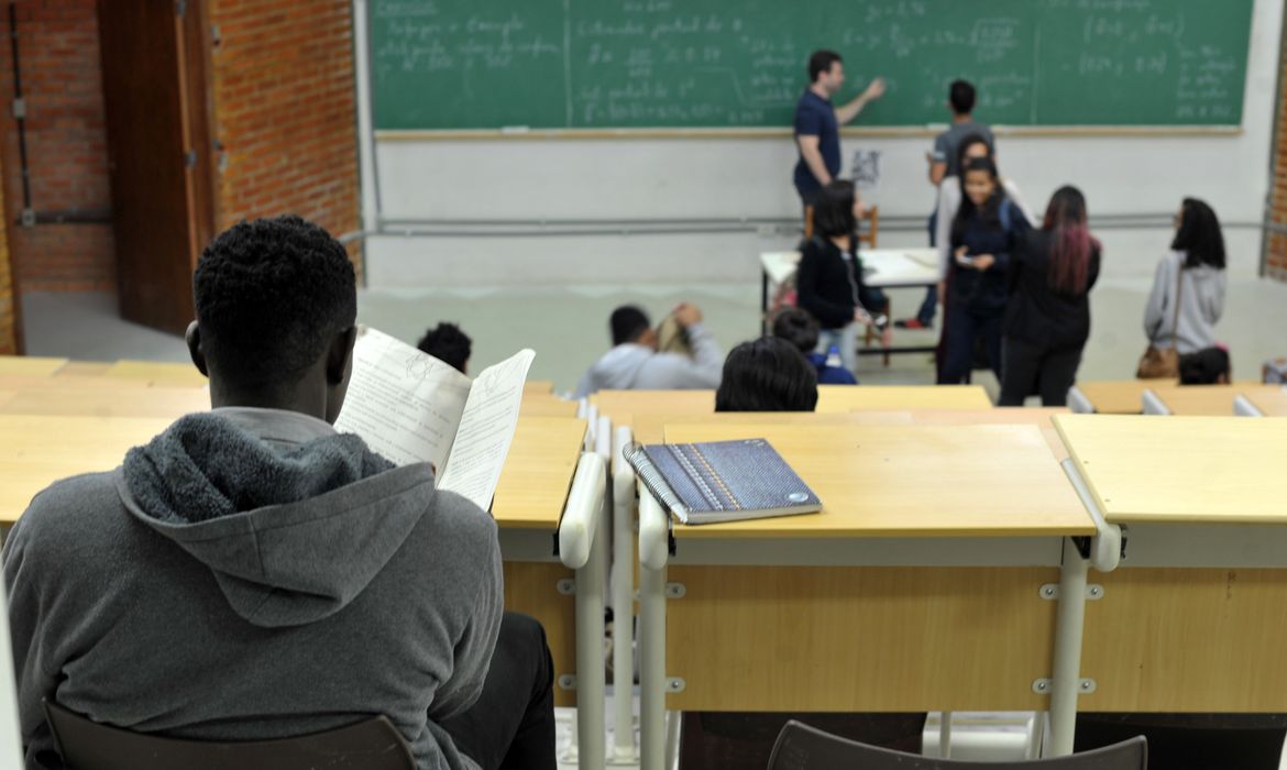 Fotografia de uma sala de uma universidade. No primeiro plano, à esquerda, um aluno negro, de costas, sentado, lendo um material durante a aula